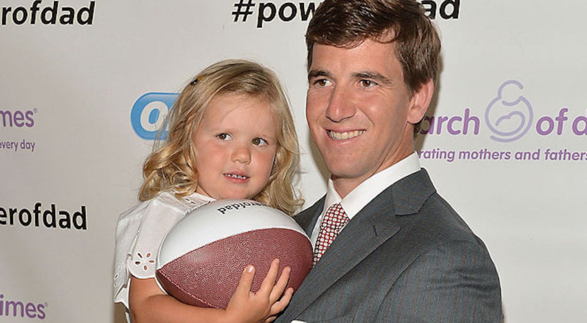 Eli Manning holding daughter Ava Francis while she holds a football and smiles for cameras.