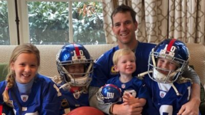 Eli Manning on the couch with his four children, all smiling at the camera and wearing his Giants jersey.