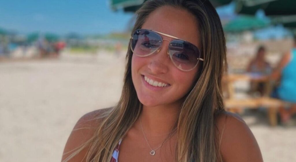 Amy Paternoster at the beach smiling at the camera in an american flag bikini and aviator sunglasses.