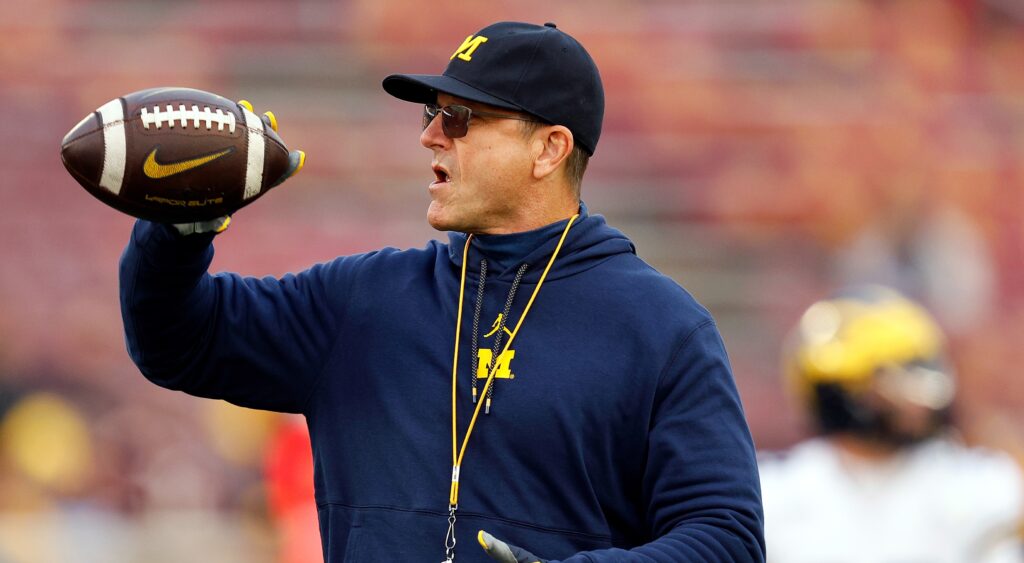 Jim Harbaugh of Michigan Wolverines holding football while looking on.