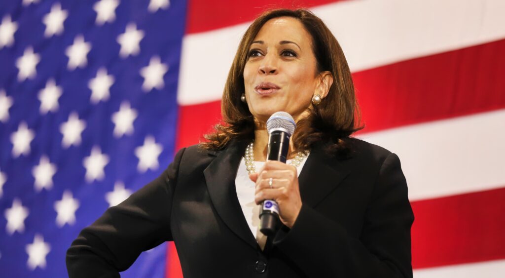 Kamala Harris speaking at an event with the USA flag in the background. Nick Saban is being named as her possible VP.